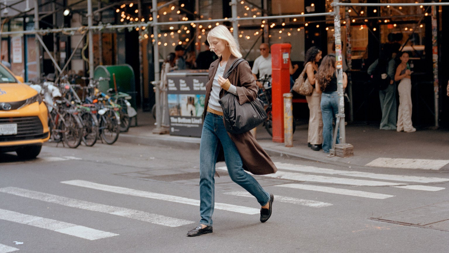 Woman crossing a street in an urban setting with a taxi and pedestrians in the background holding a black TYVY tote bag.