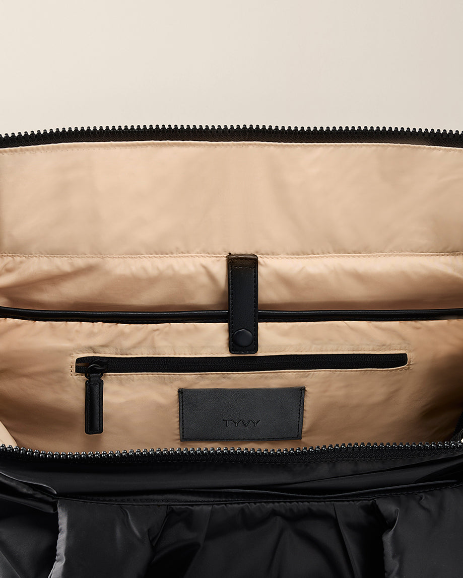 Close-up of a open totebag with black zipper and branding on a beige background. TYVY Aviator Tote bag optimized for travel.
