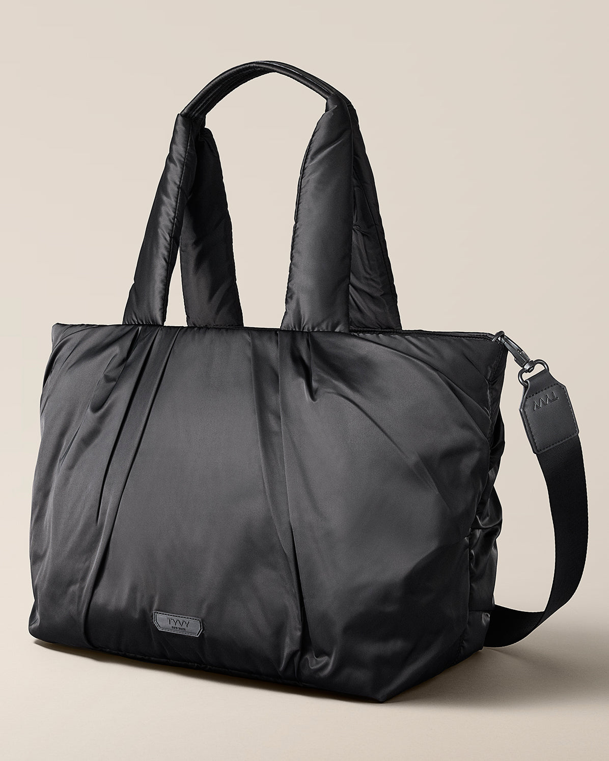 Black tote bag with a shoulder strap on a beige background, TYVY Aviator Tote bag optimized for travel.