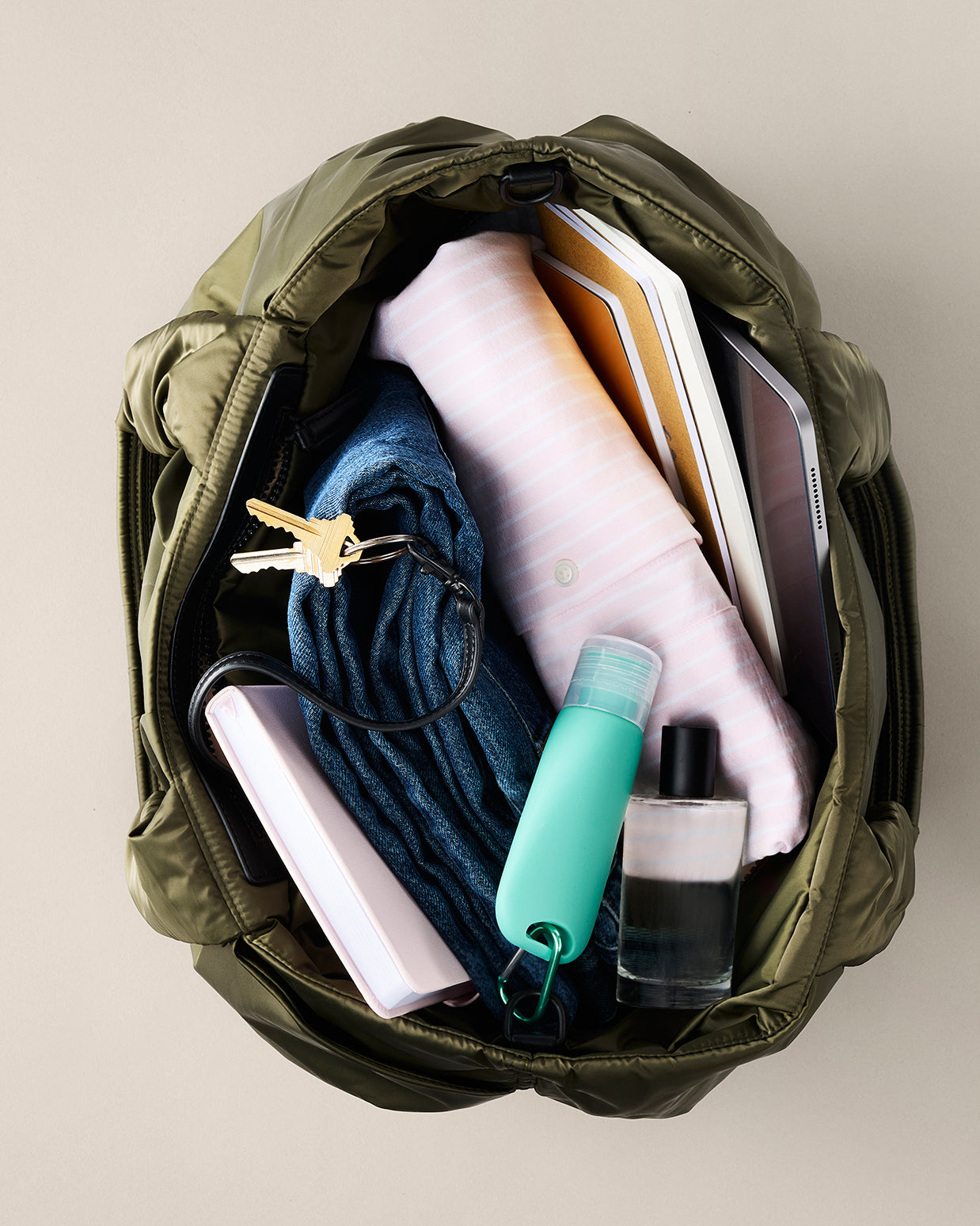 Open green backpack with various items including a phone, keys, and bottles on a beige background. TYVY Aviator Tote bag optimized for travel.