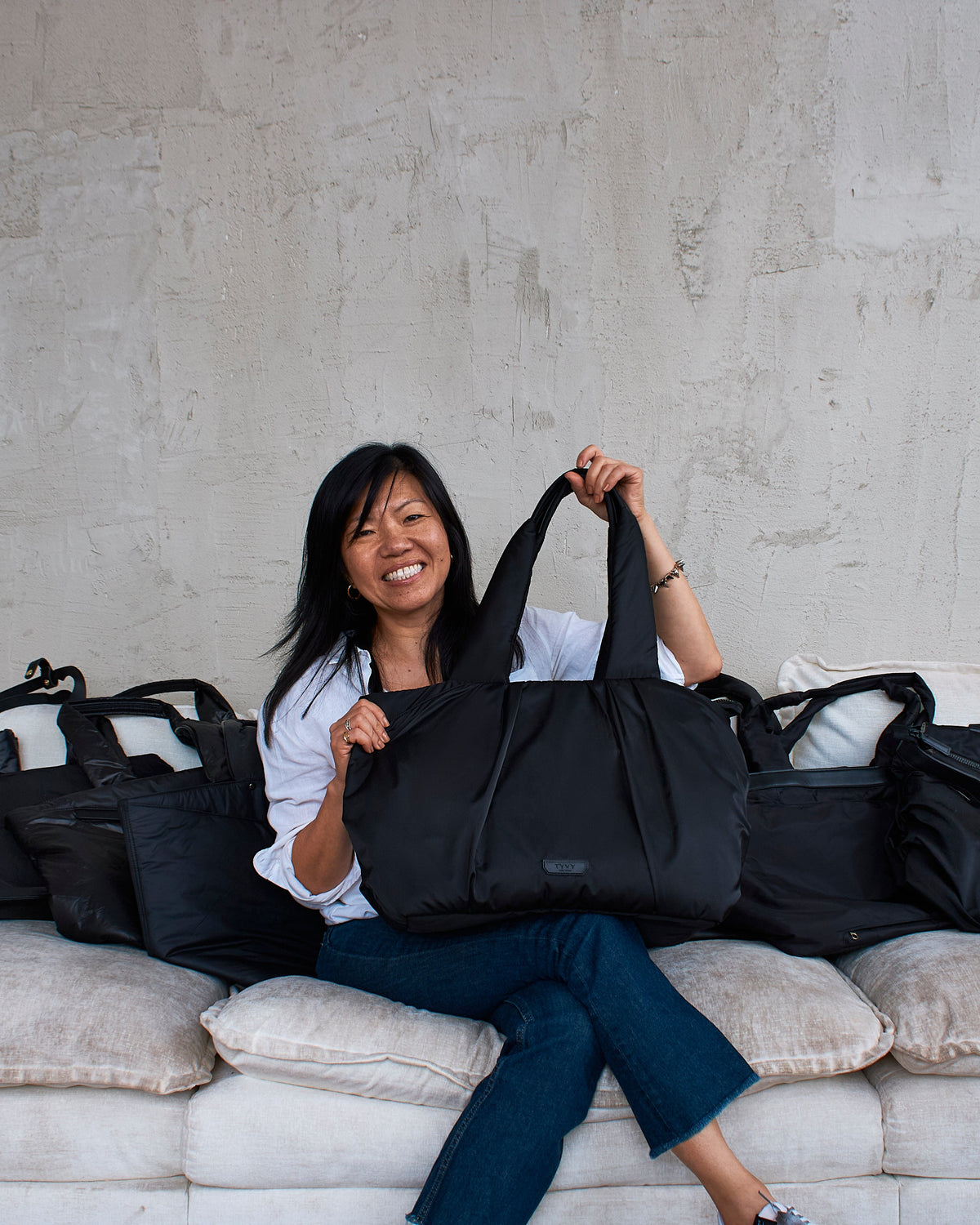 Tina Yau sitting on a couch with multiple black bags around her, against a plain wall.