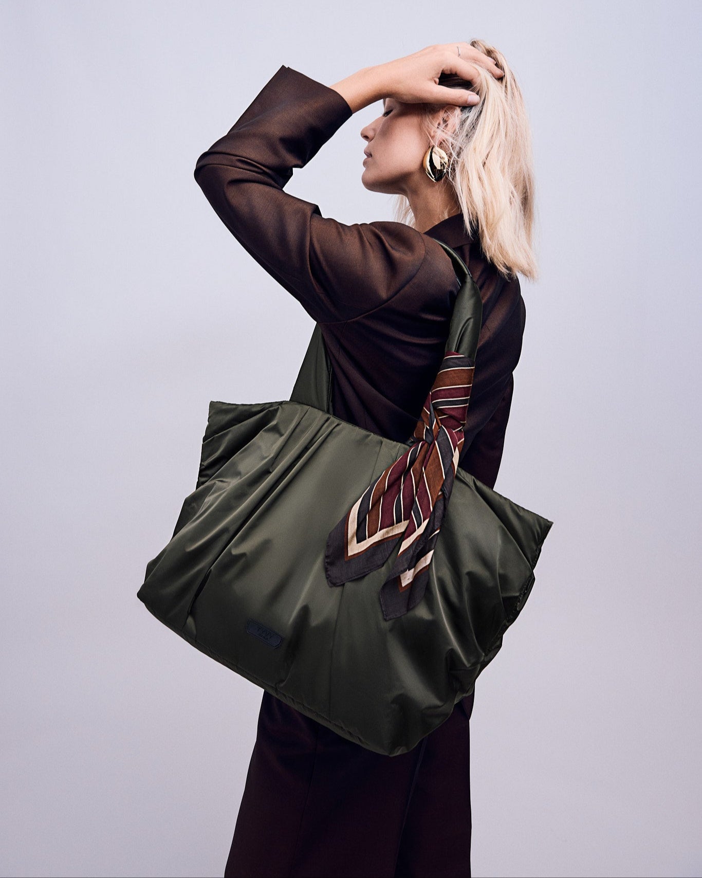 Woman holding a large olive green TYVY tote bag with a patterned tie over her shoulder against a light gray background