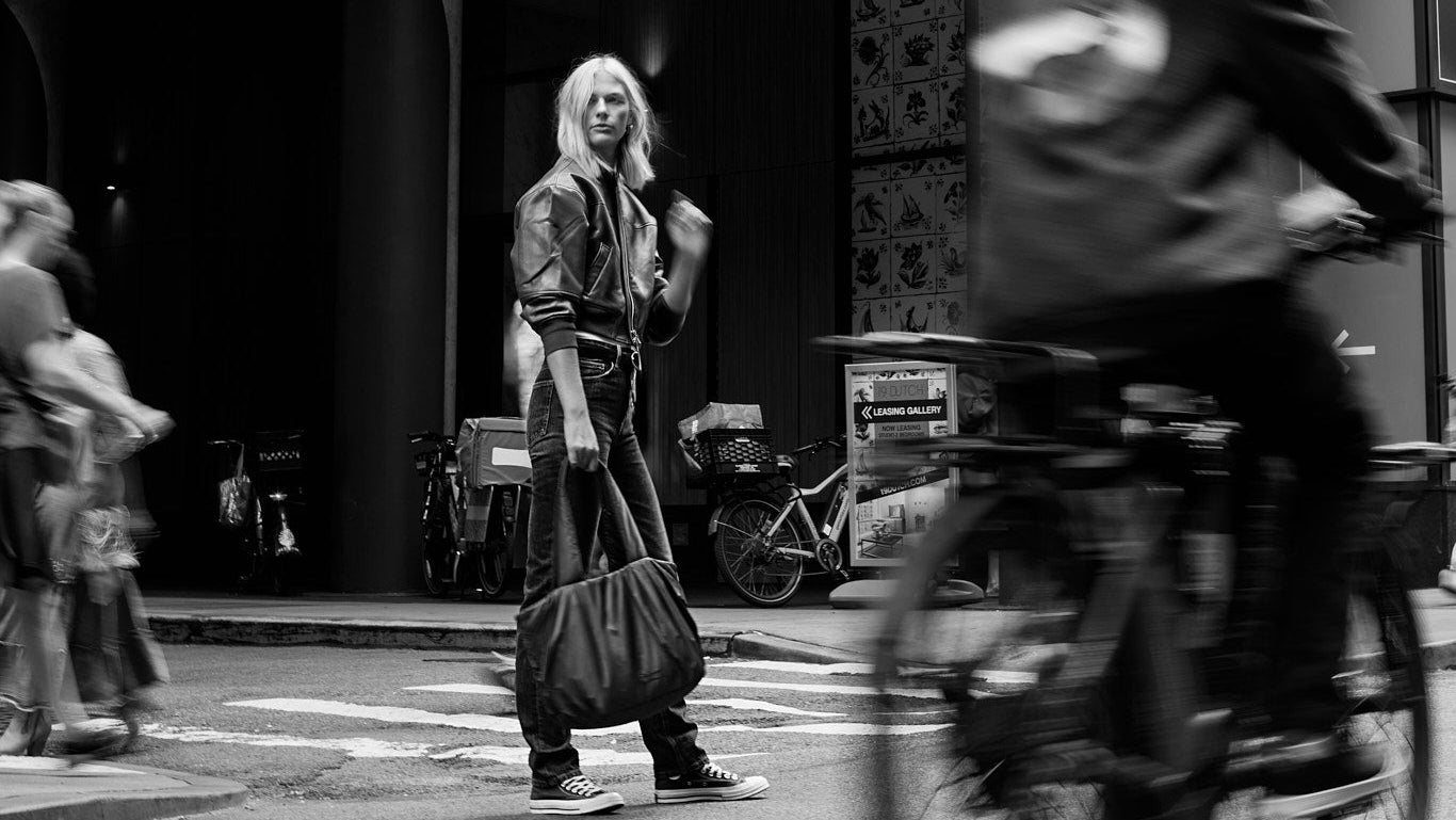 Black and white street scene with a person on a bicycle and a woman standing in front of a building holding a black TYVY tote bag.